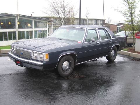 1991 Ford LTD Crown Victoria - dark navy Ford LTD Crown Victoria four-door sedan, largely unmarked, parked on a wet lot in front of a building.