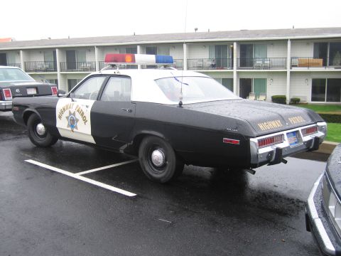1991 Ford LTD Crown Victoria - black-and-white Highway Patrol Plymouth coupe with roof lightbar parked in a row of patrol cars before a motel on a wet lot.