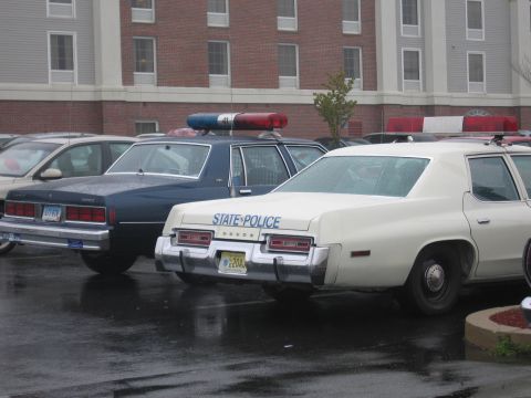1991 Ford LTD Crown Victoria - Two classic police sedans parked on a wet lot, foreground a white New Jersey State Police Dodge with roof lightbar, rear view.