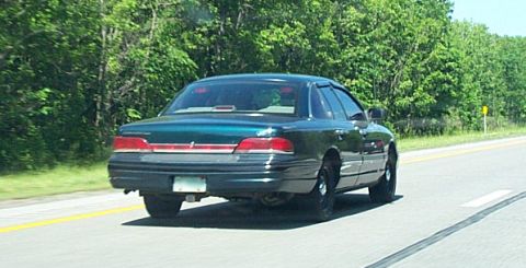 2001 Ford Crown Victoria - A dark green 2001 Ford Crown Victoria sedan is seen from the rear on a highway surrounded by trees.