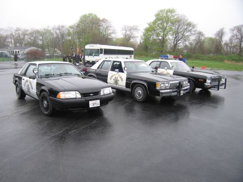 1991 Ford LTD Crown Victoria - row of black-and-white Highway Patrol cars including a Mustang and full-size sedans, with a white bus behind, on a wet lot.