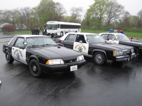 1991 Ford LTD Crown Victoria - row of black-and-white Highway Patrol patrol cars and a white bus parked on a rainy lot, front three-quarter view.