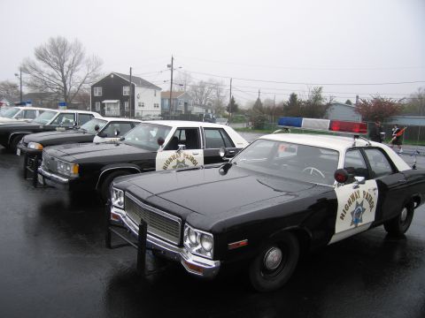 1991 Ford LTD Crown Victoria - lineup of black-and-white Highway Patrol sedans with front push bars and lightbars parked on a foggy, wet lot.
