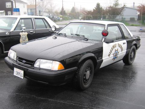 1991 Ford LTD Crown Victoria - black-and-white California Highway Patrol Ford Mustang 5.0 coupe parked on a wet lot among other classic patrol cars.