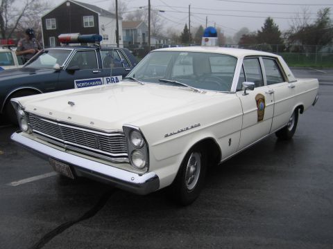 1991 Ford LTD Crown Victoria - white Ford Galaxie 500 four-door sedan outfitted as a police car with a blue roof dome light, parked on wet pavement near other cruisers.