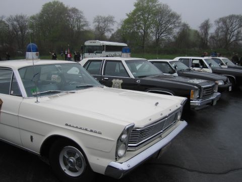 1991 Ford LTD Crown Victoria - lineup of vintage police cruisers parked side by side on wet pavement, led by a white Ford Galaxie 500 with a blue dome light.