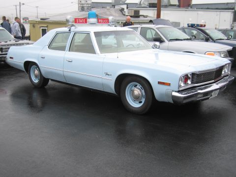 1991 Ford LTD Crown Victoria - light blue Plymouth Fury four-door sedan police car parked under a State Police sign on a wet lot among other cruisers.