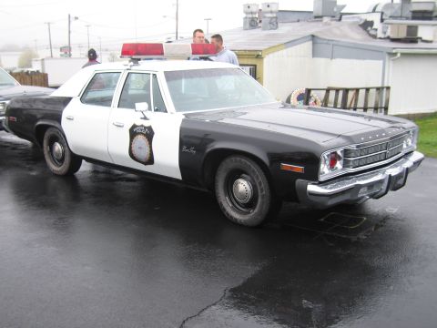 1991 Ford LTD Crown Victoria - black-and-white Plymouth Fury four-door police sedan with a roof lightbar and door badge parked on wet pavement.