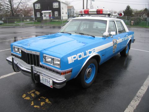 1991 Ford LTD Crown Victoria - blue-and-white Dodge police sedan lettered POLICE with NYPD-style markings and a roof lightbar, parked in a wet lot.