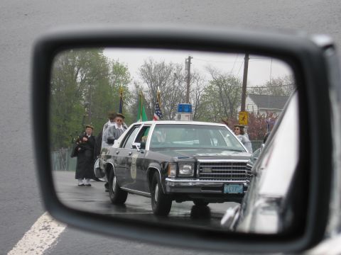 1991 Ford LTD Crown Victoria - side-mirror reflection showing a dark four-door police sedan with a dome light, onlookers and flags on a gray rainy day.
