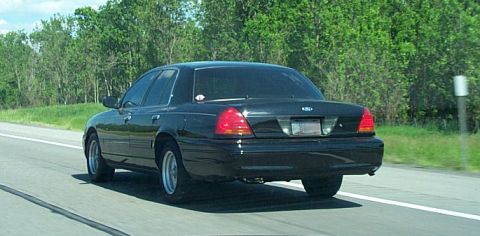 2001 Ford Crown Victoria - A black Ford Crown Victoria is captured from behind while driving on a highway with trees lining the road.