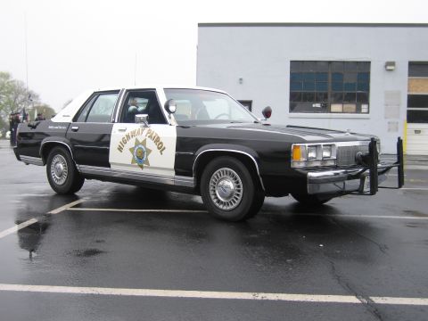 1991 Ford LTD Crown Victoria - black-and-white Ford LTD Crown Victoria California Highway Patrol cruiser with a push bar parked outside a garage building.