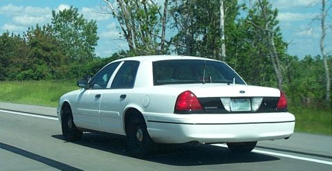 2001 Ford Crown Victoria - A white 2001 Ford Crown Victoria sedan is seen from the rear on a highway with trees in the background.