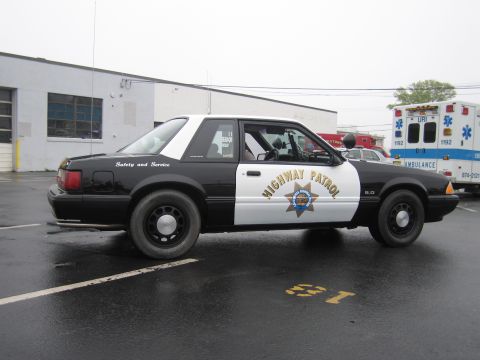1991 Ford LTD Crown Victoria - black-and-white Fox-body Ford Mustang 5.0 California Highway Patrol cruiser shown in side profile on a wet lot near an ambulance.