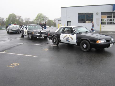 1991 Ford LTD Crown Victoria - Black-and-white CHP Ford Mustang LX police cruiser parked on a wet lot alongside older Crown Victoria patrol cars.