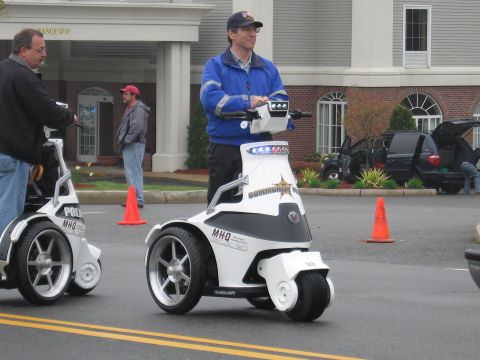 1991 Ford LTD Crown Victoria - man in a blue jacket rides a white three-wheeled stand-up police personal mobility vehicle past traffic cones.