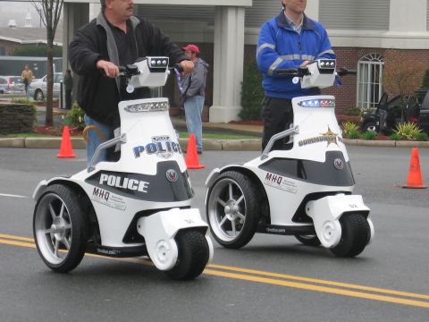 1991 Ford LTD Crown Victoria - Two men ride white three-wheeled stand-up police personal mobility vehicles side by side near orange cones.