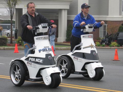 1991 Ford LTD Crown Victoria - Two men ride white three-wheeled stand-up police personal mobility vehicles marked Police on a parking lot.