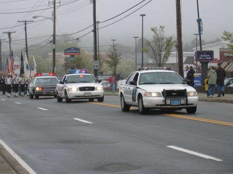 1991 Ford LTD Crown Victoria - White police cruisers with flashing lights lead marchers down a foggy street past strip-mall storefronts.