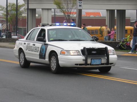 1991 Ford LTD Crown Victoria - White Ford Crown Victoria police cruiser with push bar and blue lights drives past a school bus and onlookers.