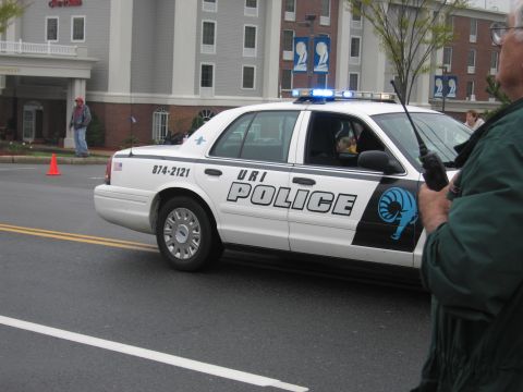 1991 Ford LTD Crown Victoria - White URI Police Ford Crown Victoria cruiser with a blue ram logo, viewed past an officer holding a radio.