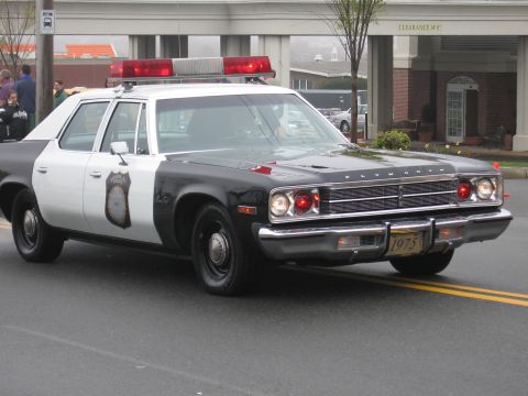 1991 Ford LTD Crown Victoria - Black-and-white 1975 Plymouth police sedan with dome lights and a 1975 license plate driving on a street.