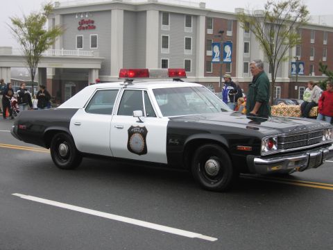1991 Ford LTD Crown Victoria - Side three-quarter view of a black-and-white vintage Plymouth police sedan with red dome lights in a parade.