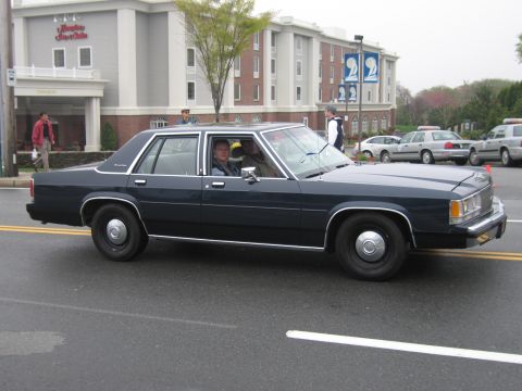 1991 Ford LTD Crown Victoria - dark navy Ford LTD Crown Victoria four-door sedan with a driver inside cruises down a street past a hotel and parked cars.