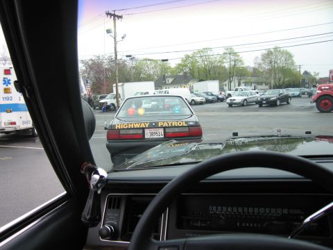 1991 Ford LTD Crown Victoria - Looking through a windshield over the dashboard at a black Highway Patrol cruiser stopped ahead, license plate 389514, in a parking lot.