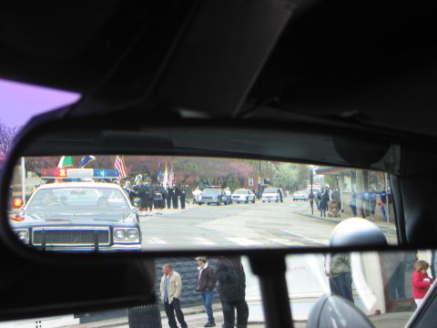 1991 Ford LTD Crown Victoria - car's rearview mirror reflects a black police cruiser, flags, parked patrol cars and pedestrians along a town street.