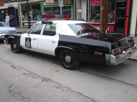 1991 Ford LTD Crown Victoria - black-and-white 1970s police sedan with a rooftop beacon sits parked curbside in front of a VCR repair shop, rear three-quarter view.