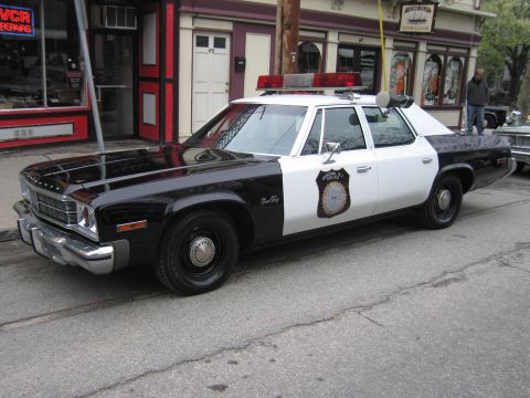 1991 Ford LTD Crown Victoria - Front three-quarter view of a black-and-white mid-1970s police sedan with a gold star-badge door decal parked in front of a repair shop.