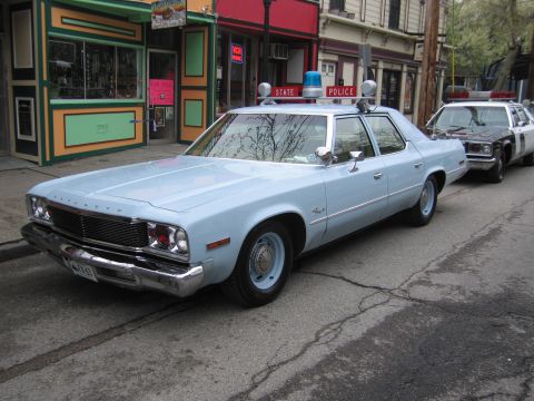 1991 Ford LTD Crown Victoria - light blue 1970s Plymouth four-door State Police sedan with red-and-blue rooftop lights parked curbside on a town street.