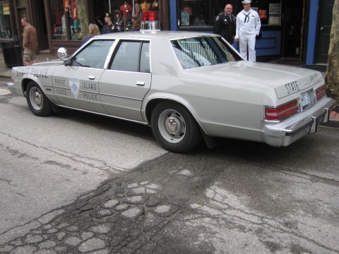1991 Ford LTD Crown Victoria - Rear three-quarter view of a silver Rhode Island State Police four-door sedan parked curbside with uniformed officers standing nearby.