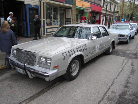 1991 Ford LTD Crown Victoria - Front three-quarter view of a silver Chrysler-style State Police sedan parked curbside, with more cruisers lined up behind it.