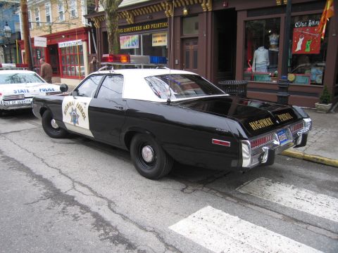 1991 Ford LTD Crown Victoria - Rear three-quarter view of a black-and-white Highway Patrol sedan with a gold star-badge door decal parked curbside near storefronts.