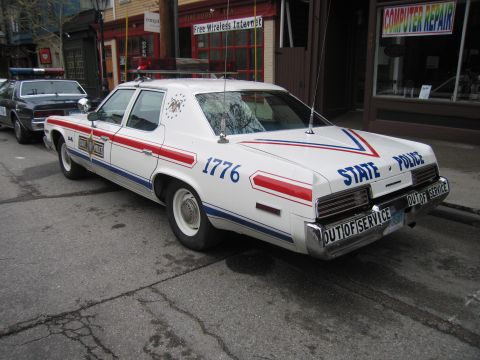 1991 Ford LTD Crown Victoria - Rear three-quarter view of a white State Police sedan with red-and-blue stripes, numbered 1776, marked Out of Service, parked curbside.