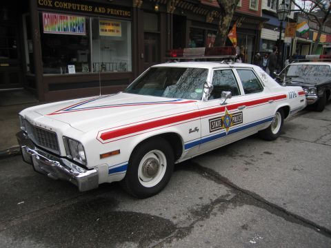 1991 Ford LTD Crown Victoria - Front three-quarter view of a white State Police sedan with red-and-blue stripes and a star-badge door decal parked on a town street.