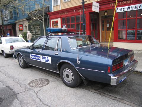 1991 Ford LTD Crown Victoria - Rear three-quarter view of a dark blue North Haven PBA police sedan with a rooftop light bar parked curbside outside a coffee shop.