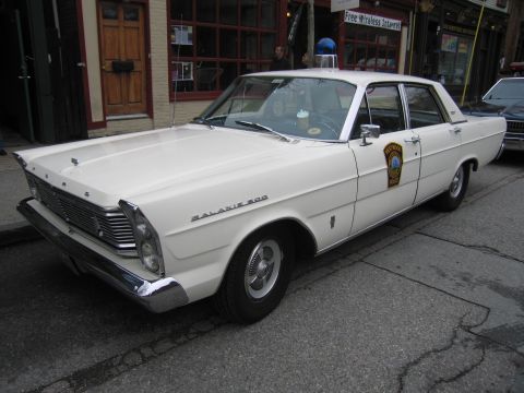 1991 Ford LTD Crown Victoria - Front three-quarter view of a white Ford Galaxie 500 police sedan with a blue rooftop beacon and badge decal parked curbside.