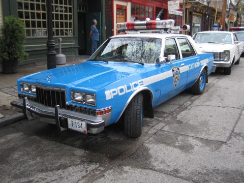 1991 Ford LTD Crown Victoria - Front three-quarter view of a blue-and-white NYPD police sedan with a rooftop light bar parked curbside on a town street.
