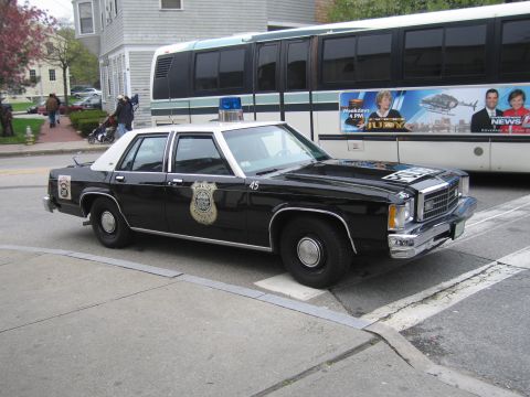 1991 Ford LTD Crown Victoria - black-and-white Ford LTD Crown Victoria police sedan numbered 45 with a badge decal parked at a curb beside a coach bus.