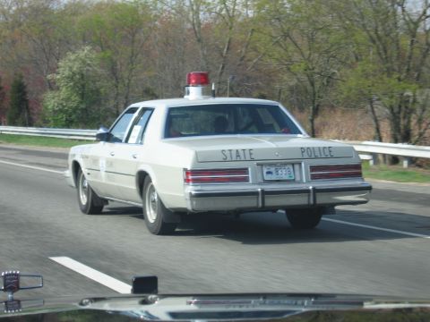 1991 Ford LTD Crown Victoria - cream-colored State Police sedan with a single red rooftop beacon drives along a tree-lined highway, seen from behind.