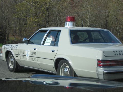 1991 Ford LTD Crown Victoria - cream Rhode Island State Police sedan with a red rooftop beacon, marked Out of Service, drives along a road, side rear view.