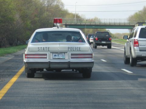 1991 Ford LTD Crown Victoria - Rear view of a cream-colored State Police sedan with a red rooftop beacon traveling on a multi-lane highway near an overpass.