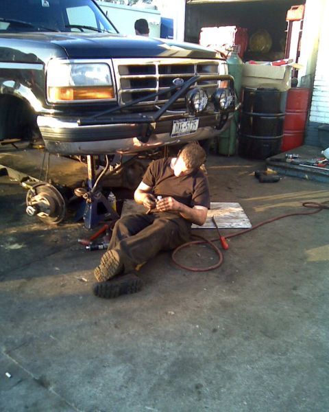 1995 Ford Bronco - A mechanic works on a lifted Ford truck in a garage setting, with various tools and parts around.