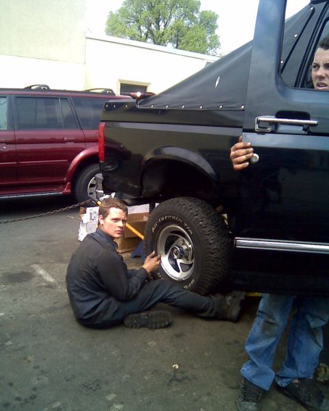 1995 Ford Bronco - A man works on the rear tire of a black SUV while another person looks out the window, with a red SUV in the background.