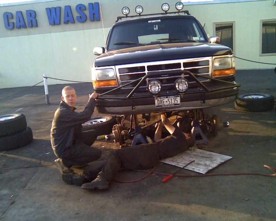 Black Ford truck on jack stands with roof light bar as two people work underneath