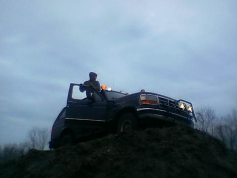 1995 Ford Bronco - A person is standing on top of a dark-colored Ford F-Series truck, which is positioned on a dirt hill.
