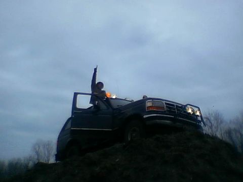 1995 Ford Bronco - A person stands atop a dark blue Ford Bronco, which is positioned on a dirt mound against a cloudy sky.